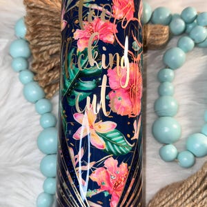 May include: A stainless steel tumbler with a floral design in pink, orange, and green on a navy blue background. The words "too f*cking cute" are written in gold script. The tumbler has a gold geometric design at the bottom.