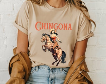 May include: A beige t-shirt with a graphic print of a woman riding a horse. The text "CHINGONA" is printed in orange above the graphic.