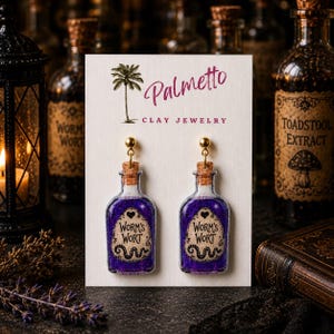 May include: A pair of dangle earrings shaped like miniature glass bottles filled with purple glitter. Each bottle features a cork stopper, a gold-tone metal hook, and a label reading Worm's Wort with a heart and snake illustration on a white display card.