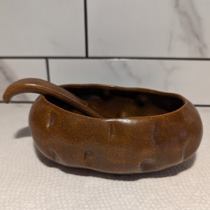 May include: A brown ceramic bowl shaped like a potato with a matching brown ceramic spoon. The bowl has a textured surface and the spoon has a curved handle.
