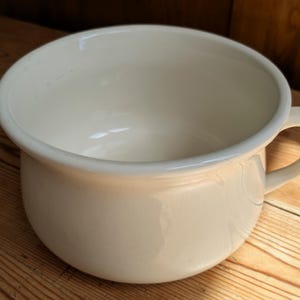 May include: A cream-coloured ceramic chamber pot with a wide, rounded basin and a single side handle. The vessel features a smooth, glossy finish and a thick, rolled rim, presenting a simple, functional design with a deep, open interior space.