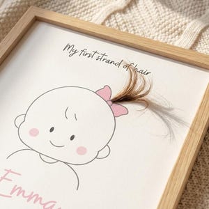 Puede incluir: Impresión enmarcada con un dibujo de bebé con un lazo rosa y el texto "My first strand of hair". Se adjunta un pequeño mechón de pelo castaño. El marco es de madera clara.