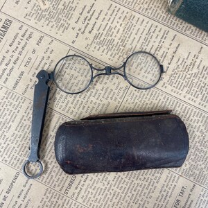 May include: Antique round eyeglasses with a folding handle and a leather case. The glasses are black and the case is dark brown, resting on a newspaper.
