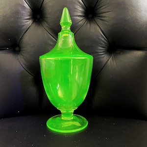 May include: A vibrant neon green glass jar with a matching pointed lid. The vessel features a smooth, translucent finish, a rounded body, a short pedestal base, and a tall, tapered finial top, creating a distinct silhouette in bright lime glass.