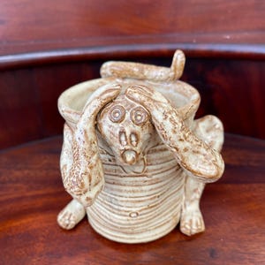 May include: A ceramic monkey figurine with its hands covering its eyes. The monkey is sitting on a wooden surface. The figurine is glazed in a light brown color with a textured finish.