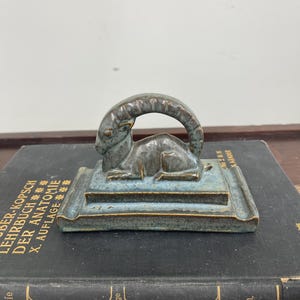 May include: A bronze-colored sculpture of a ram resting on a rectangular base with a turquoise-blue glaze. The sculpture is placed on top of a dark book with gold lettering. The book's title is partially visible.