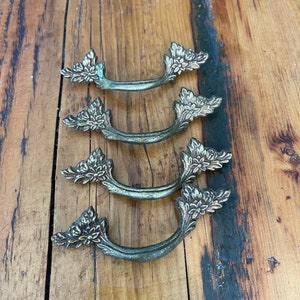 May include: Four antique brass drawer pulls with floral detailing. The pulls have a curved handle design and are arranged on a wooden surface. The brass has a slightly aged patina, adding to their vintage charm. These pulls are ideal for furniture restoration projects.