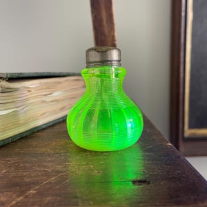 May include: A small, vintage, neon green glass bottle with a ribbed design. The bottle has a silver-colored metal cap. The bottle is sitting on a wooden surface. The bottle is approximately 3 inches tall.