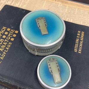 May include: Two antique powder compacts with a blue and turquoise enamel design. The compacts have a silver-colored metal rim and a rectangular silver-colored design on the lid. The compacts are on top of a dark blue book.