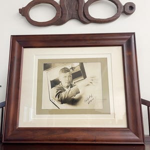 May include: A framed black and white photograph of a man looking out of a window, with a dark wooden frame. Above the frame is a decorative wooden piece with three circular openings. The photograph is signed.