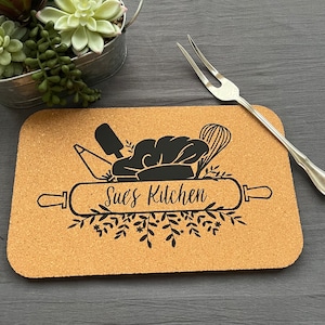 May include: A cork trivet with a black design of a rolling pin, chef's hat, whisk, and other kitchen tools. The text "Sue's Kitchen" is printed on the rolling pin.