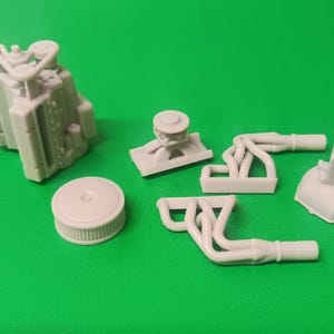 May include: A collection of light gray resin model engine parts including a V8 engine block, two exhaust headers, a circular air cleaner cover, a transmission housing, and a small mounting bracket, all displayed as separate unpainted components.