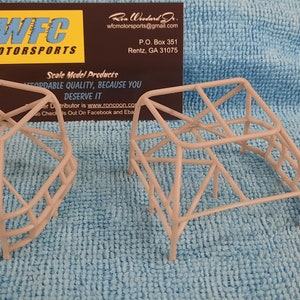 May include: Two grey metal roll cages for a model car. The cages are made of thin metal tubing and have a simple design. The cages are sitting on a blue fabric surface.
