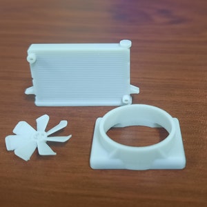 May include: Three white plastic parts for a model car engine. The parts include a radiator, a fan, and a fan housing.