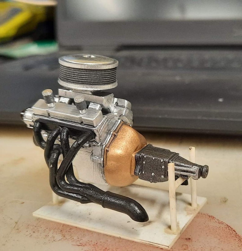 May include: A detailed model of a silver and black engine with a copper-coloured exhaust manifold. The engine is mounted on a white display stand.