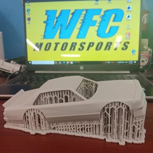 May include: A light gray 3D-printed model of a classic car. The model is detailed with open framework supporting the body. The text "WFC MOTORSPORTS" is visible on a computer screen in the background.