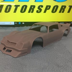May include: A tan-colored model car body, likely for a classic muscle car, with a detailed front grille and aerodynamic design. The word "MOTORSPORTS" is visible on a computer screen in the background.