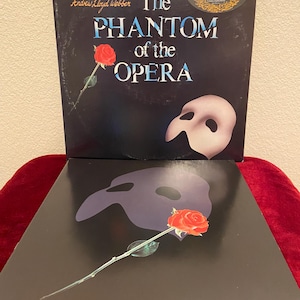 May include: Two vinyl records of the musical "The Phantom of the Opera." The top record features the title in blue and white text, a white mask, and a red rose. The bottom record shows a mask and a rose.