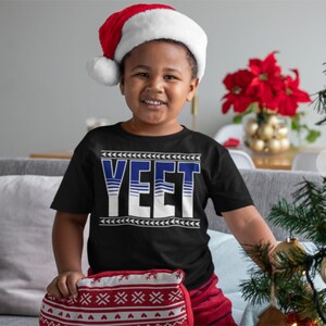 May include: A young person wearing a black t-shirt with the word "YEET" printed in white with blue stripes. The person is wearing a red Santa hat and is sitting on a couch with a red and white patterned pillow.