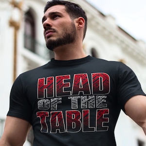 May include: Black t-shirt with the text "HEAD OF THE TABLE" in red and white. The text has a tribal design. The shirt is short-sleeved and has a crew neck.