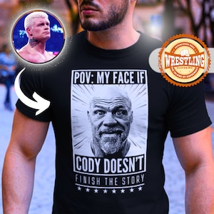 Puede incluir: Una camiseta negra con un gráfico blanco de la cara de un hombre y el texto "POV: MY FACE IF CODY DOESN'T FINISH THE STORY" con cinco estrellas debajo del texto. El gráfico está rodeado por un círculo blanco con un contorno negro. El texto "ESTABLISHED 2023 WRESTLING SUPREME THE ISLAND OF SUPREMACY" está en un círculo rojo con un contorno blanco.