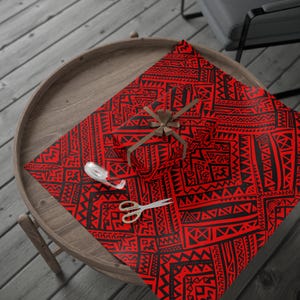 May include: A wrapped gift with a brown ribbon and a roll of white tape sits on red wrapping paper with a black tribal pattern. A pair of silver scissors is also on the paper, ready for use. The items are on a wooden table.