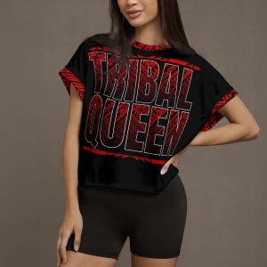 May include: Black crop top with the text "TRIBAL QUEEN" in white outlined in red. The shirt has red tribal pattern accents on the sleeves and neckline. The model is wearing black shorts.
