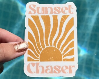 May include: A white sticker with the text "Sunset Chaser" in pink and orange letters. The sticker features a stylized orange sun with rays radiating outward.