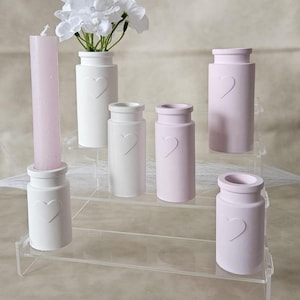 Könnte beinhalten: Ein Set aus sechs weißen und rosa Keramikvasen mit Herzmotiven, die auf einem dreistufigen, transparenten Acrylständer präsentiert werden. Die Vasen eignen sich perfekt für die Präsentation kleiner Blumensträuße oder Grüns.