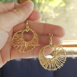 May include: A pair of gold-colored wire hoop earrings. Each earring features a circular design with a central open space, surrounded by radiating wire spokes. The earrings are held up against a blurred background.