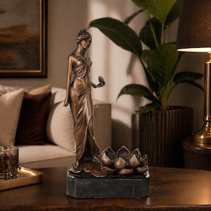 Puede incluir: Estatua de color bronce de una mujer con un vestido fluido, sosteniendo un objeto pequeño. La estatua descansa sobre una base de mármol negro sobre un diseño de flor de loto. Un acento decorativo para el hogar.