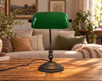 Könnte beinhalten: Eine Bankerlampe mit einem glänzenden smaragdgrünen Glasschirm und einem dunkel bronzefarbenen Metallsockel. Die Lampe verfügt über einen geschwungenen Arm, einen runden, gestuften Fuß und eine goldfarbene Zugkette, die mittig am Schirm hängt.