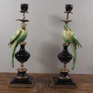 May include: Pair of decorative candle holders featuring green parrot sculptures perched atop black and gold bases. The candle holders have a black vase-shaped body and a square base with ornate gold accents. The parrots have green and yellow plumage.