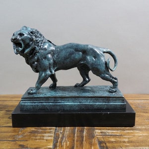 May include: A bronze sculpture of a roaring lion standing on a black marble base. The lion is depicted in a dynamic pose with its mouth open and its teeth bared. The sculpture is finished in a green patina.