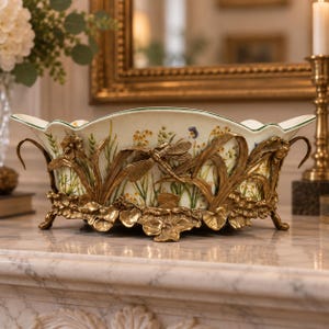 Op de afbeelding: Een ovale keramische kom met een geschulpte rand, voorzien van een geschilderd botanisch patroon van wilde bloemen en grassen. De kom is gevat in een sierlijk, antiek goudkleurig metalen frame met sculpturale libelle- en bladmotieven.