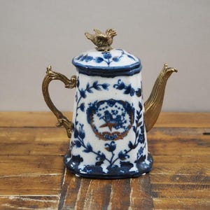 Puede incluir: Una tetera de porcelana azul y blanca con un adorno de pájaro dorado. La tetera tiene un diseño floral y está decorada con detalles dorados.