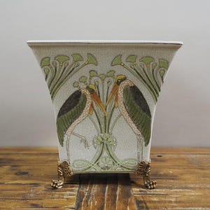 Pode incluir: Um vaso de cerâmica branco com formato quadrado e pés dourados. O vaso apresenta um design estilizado de dois pássaros com pescoços longos e folhagem verde e amarela.