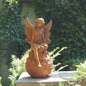 Puede incluir: Estatua de ángel de metal oxidado, de color óxido, con alas extendidas, sentada sobre una esfera texturizada. El ángel lleva un vestido fluido. La estatua está colocada sobre un fondo de follaje verde.