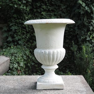 Puede incluir: Una urna blanca y ornamentada con un borde ancho y plano y una base cuadrada. La urna tiene una superficie texturizada y un diseño acanalado clásico en el cuerpo. Se encuentra sobre un fondo de exuberante follaje verde, lo que sugiere un entorno de jardín al aire libre.