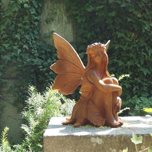 Puede incluir: Estatua de hada de color bronce con grandes alas detalladas, sentada con los brazos alrededor de las rodillas. El hada está adornada con una corona floral. La estatua está sobre una superficie de piedra, rodeada de vegetación.