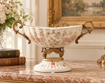 May include: A decorative porcelain bowl featuring a cream-colored base with floral patterns and a central blue bird illustration. The bowl has two ornate gold-toned metal handles and a pedestal base supported by two gold-toned swan figures.