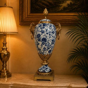 Może przedstawiać: Ozdobna porcelanowa urna z niebiesko-białym wzorem kwiatowym. Naczynie posiada złote metalowe detale, w tym uchwyty w kształcie głów baranów z pierścieniami, pokrywkę z gałką, ażurową podstawę oraz kwadratowy cokół.