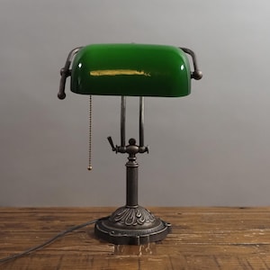 May include: A vintage-style desk lamp with a green glass shade and a bronze-colored metal base. The lamp features an adjustable shade and a pull chain. The base has an ornate, decorative design. The lamp is on a wooden surface.