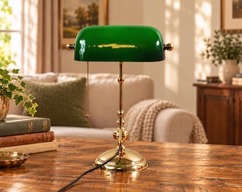 Könnte beinhalten: Eine Bankerlampe mit einem polierten goldfarbenen Metallsockel und einem zylindrischen grünen Glasschirm. Die Lampe verfügt über einen vertikalen Ständer mit dekorativen Elementen, einen runden Fuß und eine goldene Zugkette zur Bedienung.