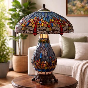 May include: A stained glass table lamp with a dragonfly design. The lamp features a colorful shade with red, yellow, blue, and orange accents. The base is a dark brown color, and the lamp sits on a round wooden table.
