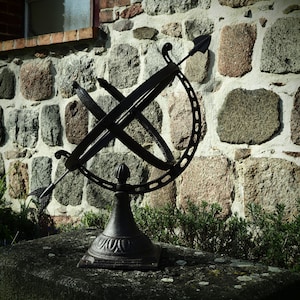 May include: A black metal sundial with an arrow pointing towards the sky, mounted on a stone base in front of a stone wall.