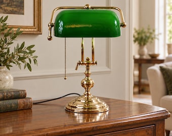 Könnte beinhalten: Eine Bankerlampe mit einem polierten Messingsockel und einem zylindrischen grünen Glasschirm. Die Lampe verfügt über einen goldfarbenen Metallrahmen, einen verstellbaren Armmechanismus und eine Kugelkette als Zugschalter an der Seite.