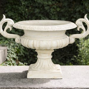 Könnte beinhalten: Cremefarbene, verzierte Urne mit dekorativen Griffen. Die Urne hat eine breite, flache Schale und steht auf einem quadratischen Sockel. Die Griffe sind geschwungen und detailliert. Die Urne besteht aus einem steinartigen Material.