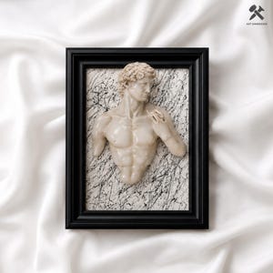 Puede incluir: Una pieza de arte mural enmarcada que presenta una escultura tridimensional de color blanco roto de un busto de torso masculino centrado sobre un fondo blanco con salpicaduras de pintura negra. El marco es negro y contiene el texto ART DIMENSION.