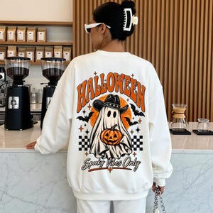 Könnte beinhalten: Ein weißes Langarm-Sweatshirt mit einem großen Grafikdruck auf der Rückseite. Das Design zeigt den Schriftzug "HALLOWEEN" und "Spooky Vibes Only" sowie einen Geist mit Cowboyhut, der einen Kürbis hält, umgeben von Fledermäusen und Sternen.